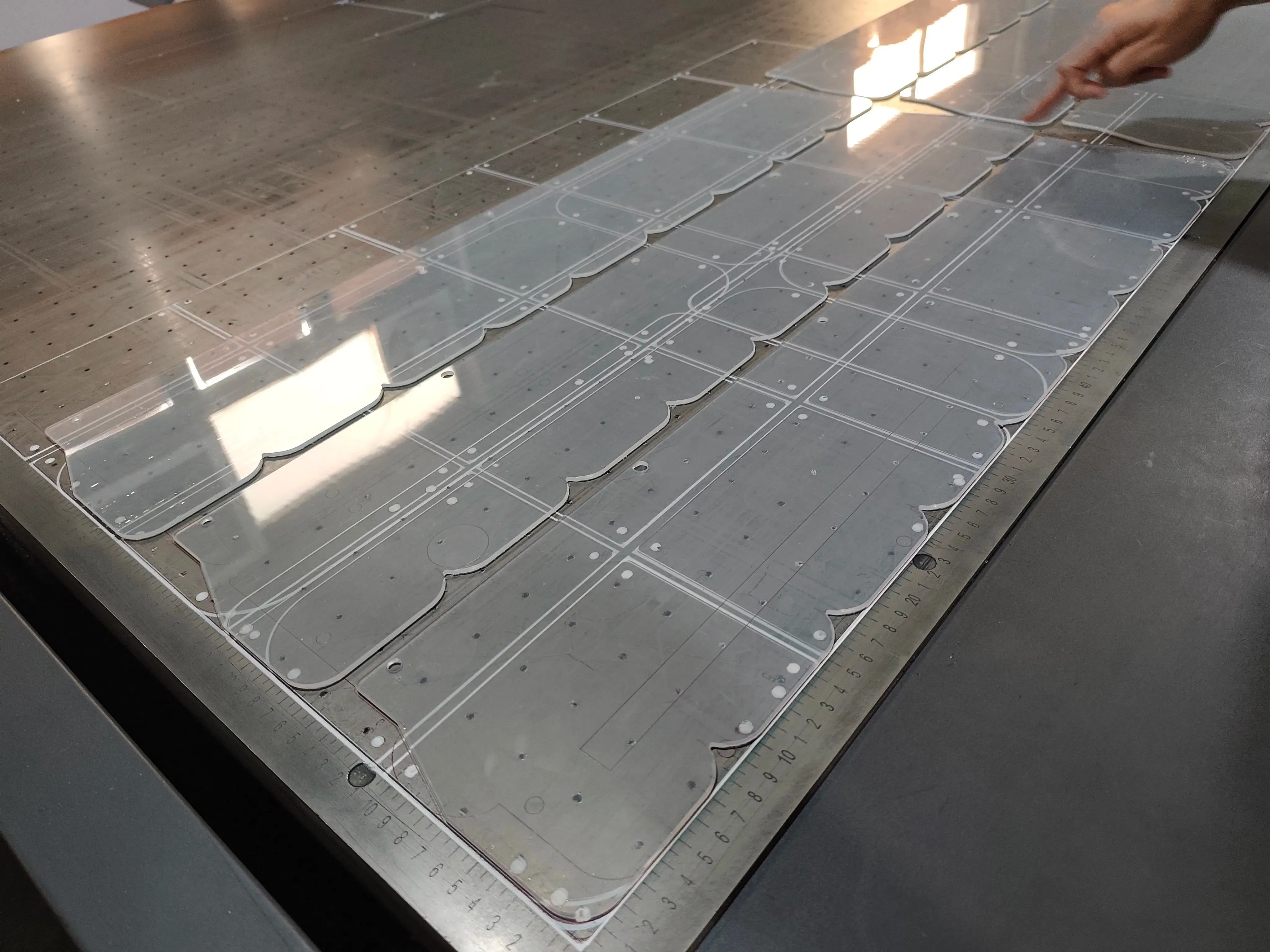 Multiple transparent acrylic substrates (game machine part blanks) placed on a UV flatbed printer platform in a factory, with a worker's finger pointing at the material—ready for UV direct printing, and the platform has a graduated ruler for positioning.