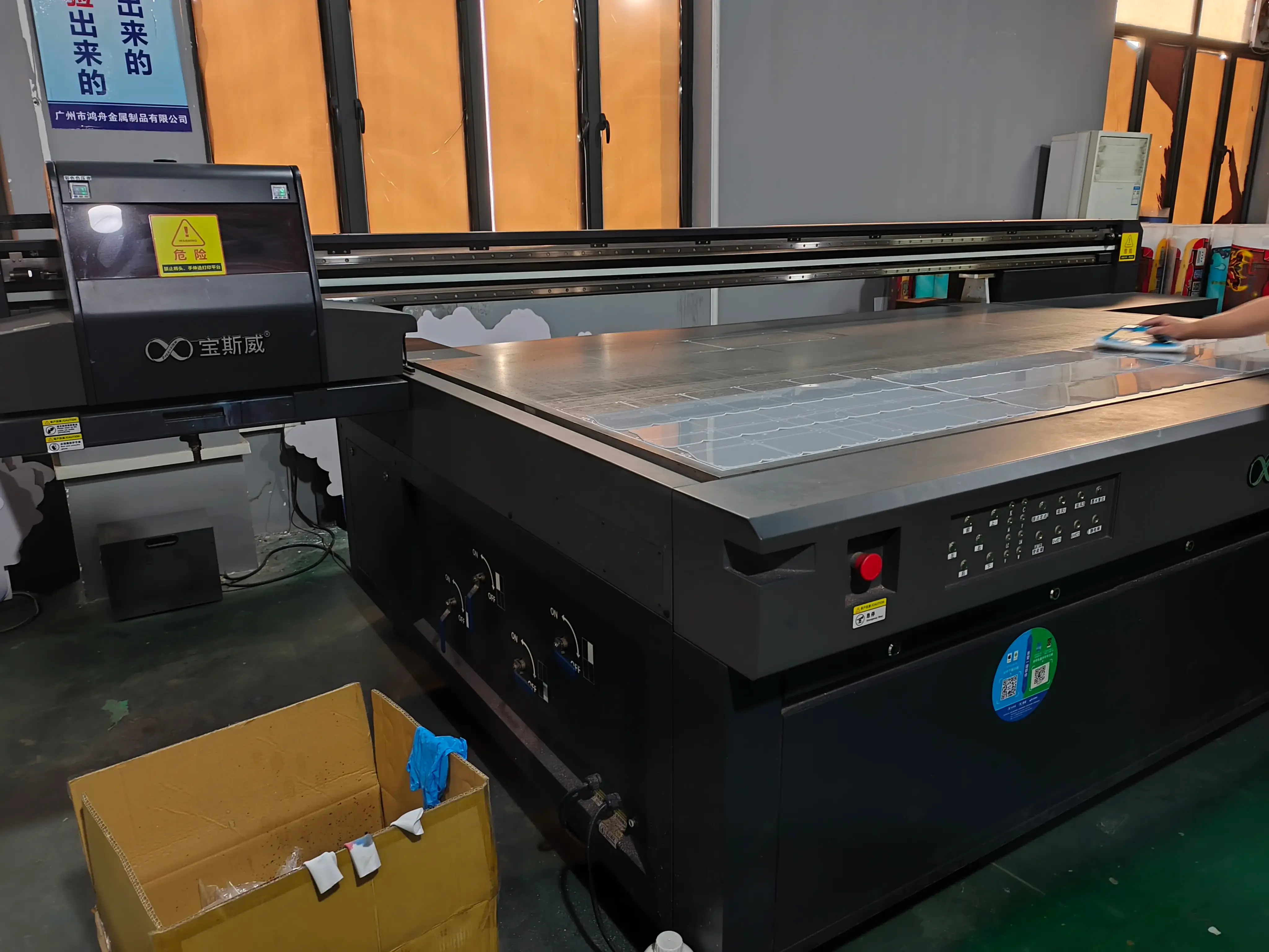 Baosiwei UV flatbed printer in a factory workshop, with a worker operating the equipment and printing materials placed on the platform—used for UV direct printing processing of materials like game machine parts.
