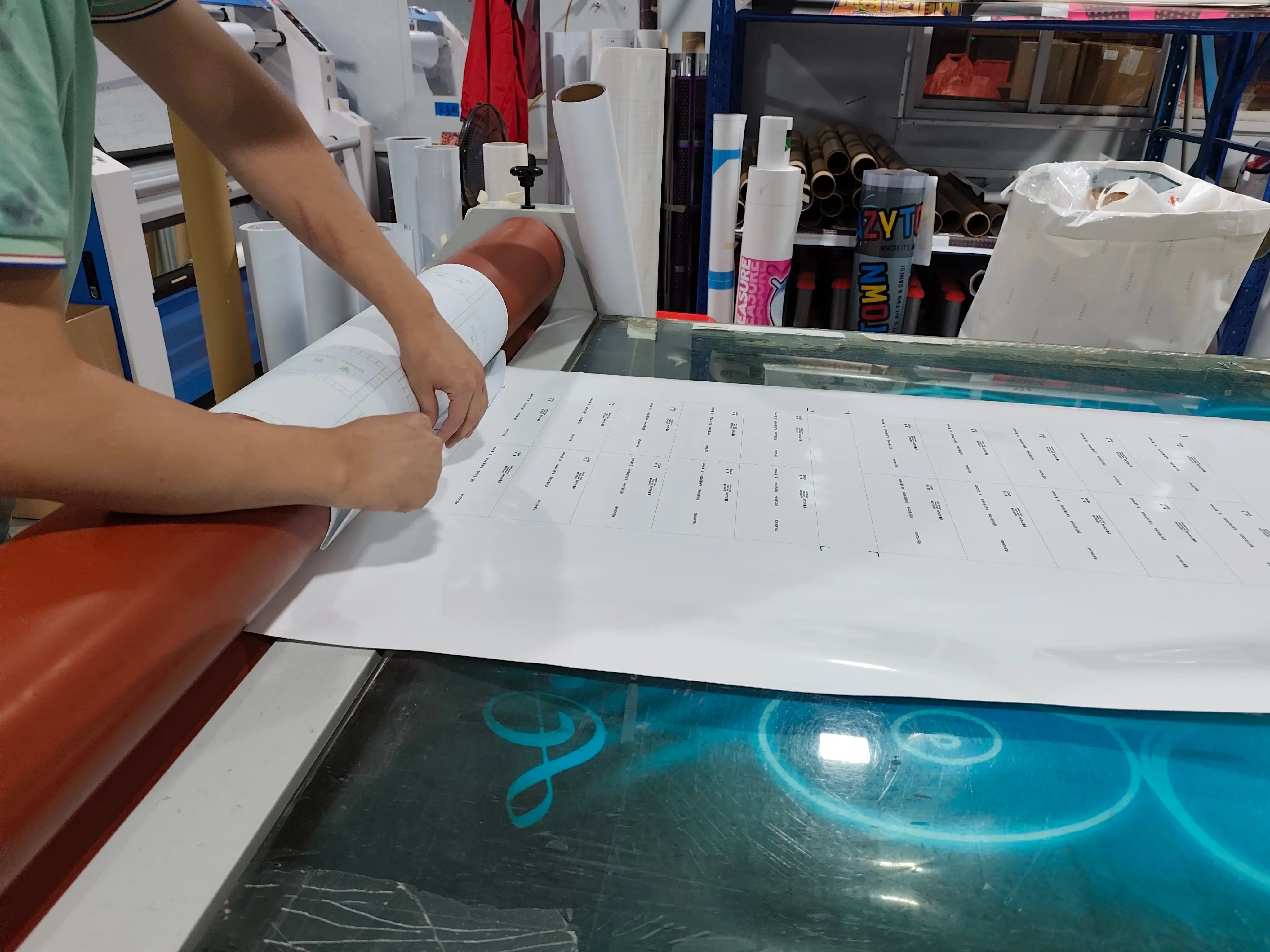 Worker laminating printed material (for game machine accessory parts) on processing equipment in print factory workshop (post-printing lamination process), with tables on the material and print supply rolls stacked in the background.