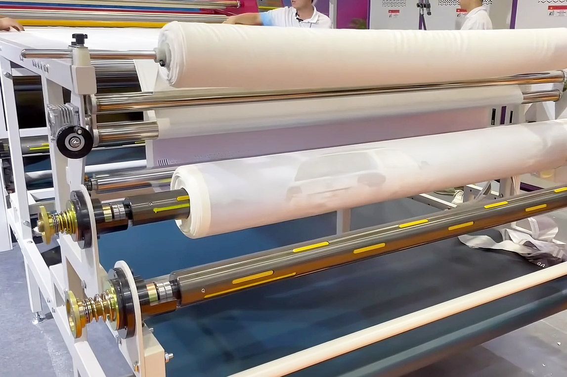 Heat transfer calender machine in action, showing fabric being conveyed through the drum for sublimation printing, producing vibrant and uniform colors for textiles, garments, and banner materials.