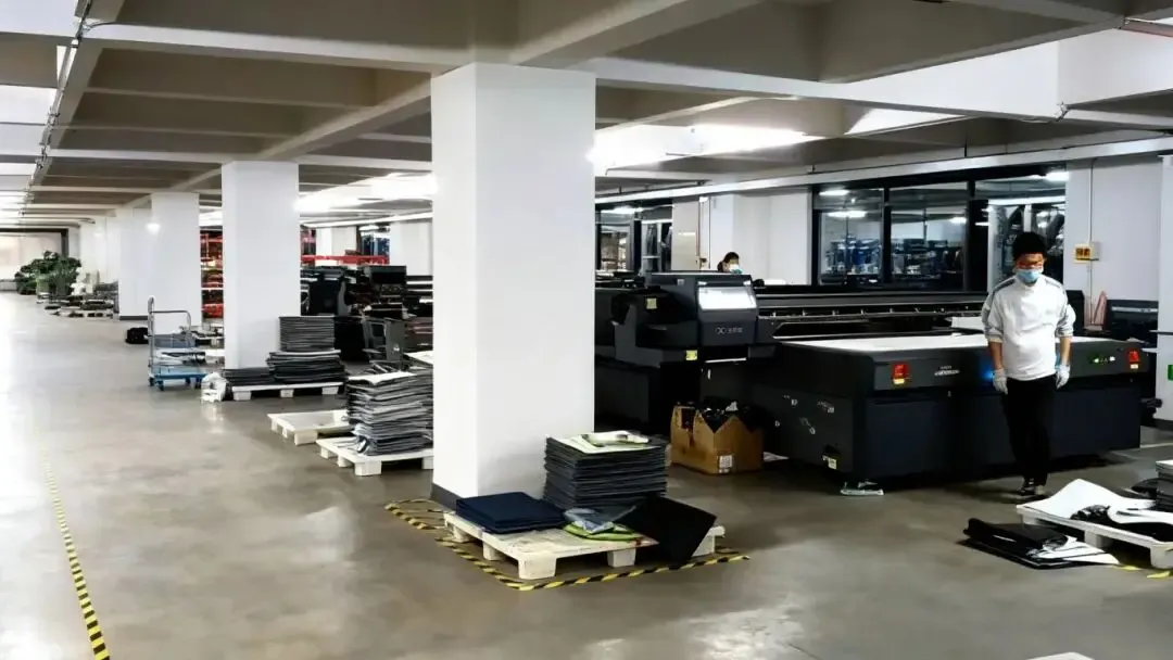 Industrial printing factory workshop equipped with Baosiwei UV flatbed printers, stacks of mat printing materials (sheets, boards) placed on pallets
