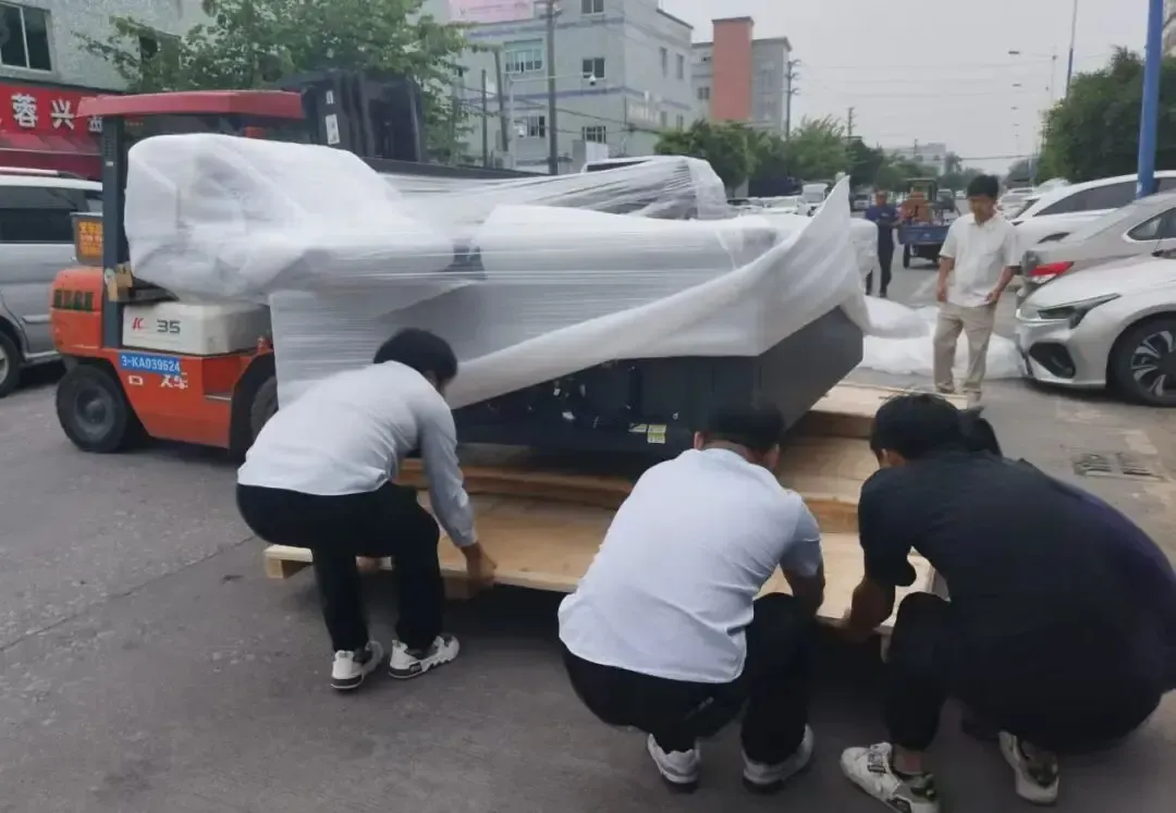 Workers coordinating with a forklift to handle a wrapped UV flatbed printer at an outdoor location; this is part of the disassembly, transportation or pre-installation process of the equipment.